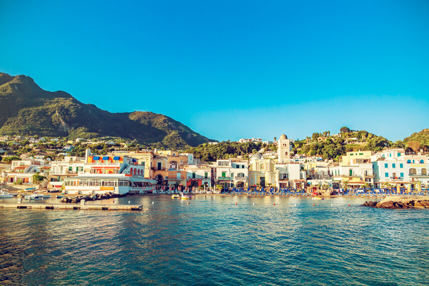 A view of Lacco Ameno, a small town on the island of Ischia, Italy. It's a warm sunny day, the sea is blue, there are sun loungers out on the small beach and the pretty pastel buildings are backed by green mountains.