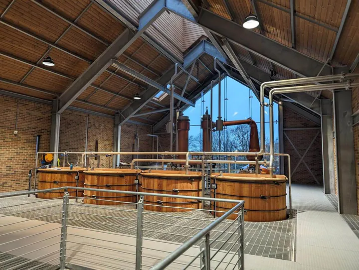 The Ad Gefrin whisky distillery. A large room with a wooden ceiling and industrial-meets-traditional vibes. Four large wooden vats are in the middle of the room, with two large copper stills at the end of the room by the window.