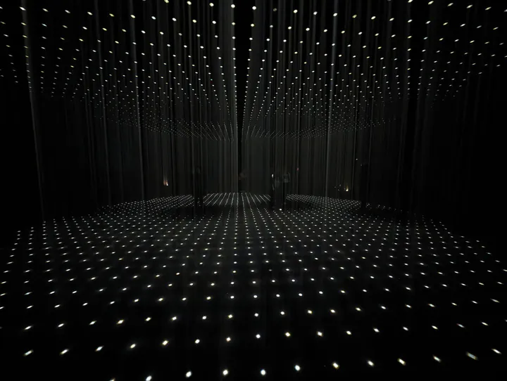 The Living Room piece by Random International. A dark room is punctuated by bars of light. Some people are stood in empty patches.