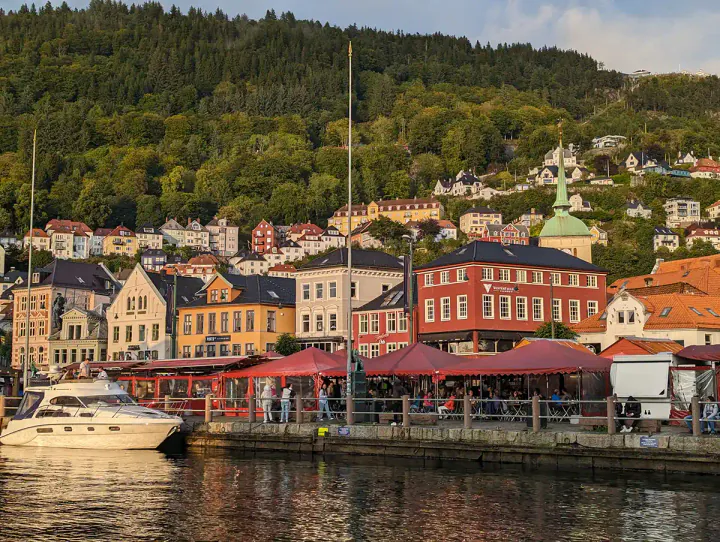 The harbour in Bergen. Colourful, low-rise buildings in red, yellow and white are clustered around a harbour. There is a thickly wooded hill, with more colourful buildings leading up the hill.