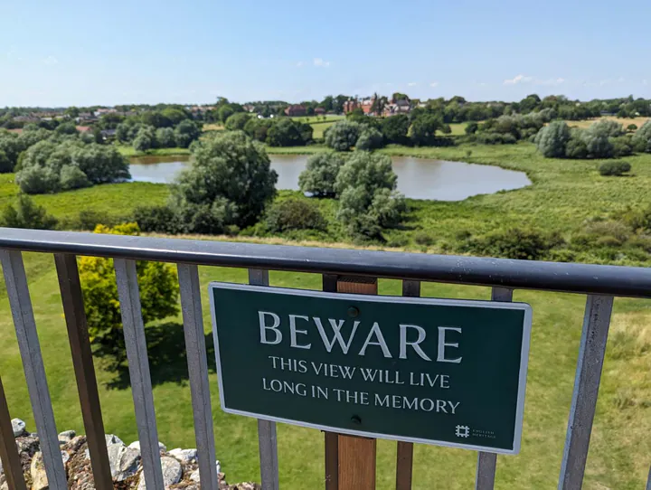 A view from Framlingham Castle's walls. A sign reads "Beware, this view will live long in the memory". The view is of green fields and a lake.