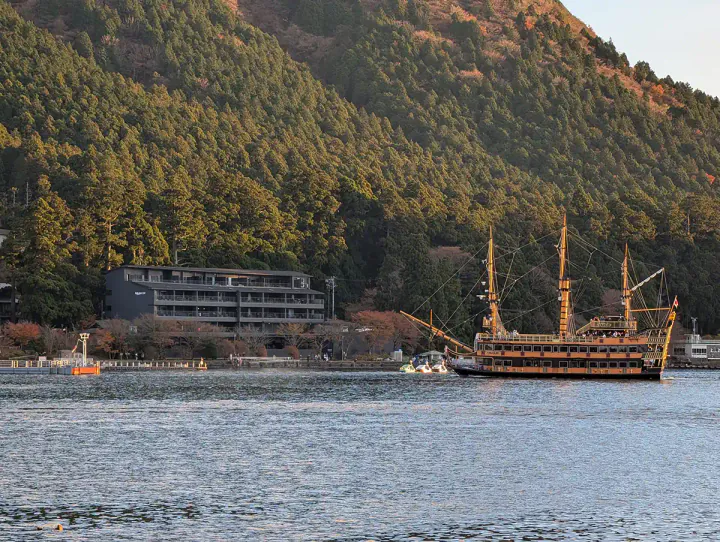 Rakuten Stay Fujimi Terrace from near the Hakone Shrine Torii gate. A dark-coloured apartment building sits between the lake and a forested hillside in Hakone, Japan. A pirate ship is crossing the lake in front of the apartment building.