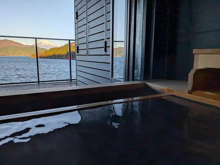 A relaxing soak in a private onsen in Hakone. The photographer is in a hot spring bath, looking out at a view of a beautiful lake and Mount Fuji.