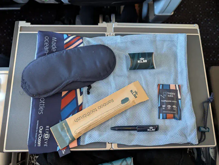 The contents of the KLM Premium Comfort amenity kit. An eye mask, earplugs, a bamboo toothbrush, a small pen and some chewable toothpaste tablets are laid out on the mesh bag they came in.