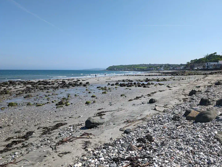 A wide sandy beach with some rocks. The sea in the background is blue and there are some green hills in the distance