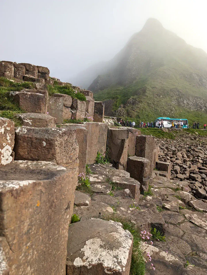 Basalt columns at the Giant's Causeway. There is a bus in the background and a spiky green cliff, which is partially shrouded in mist.