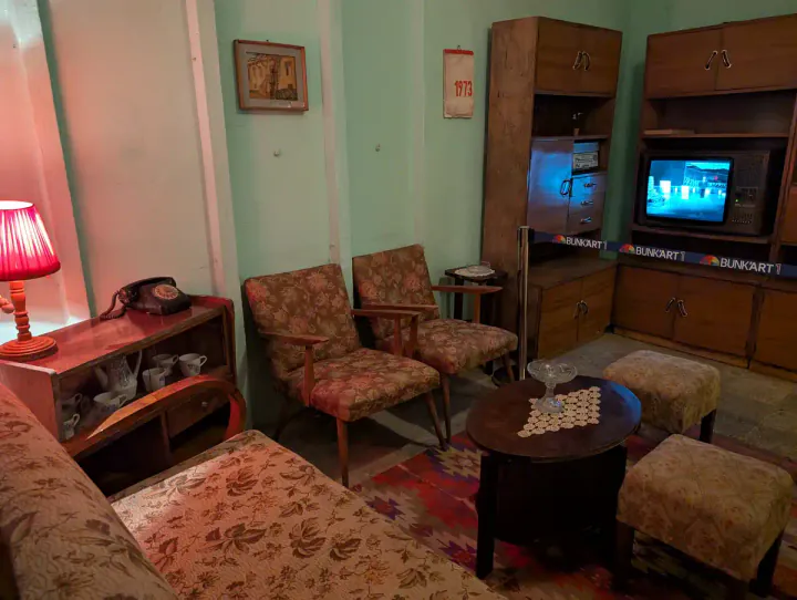 A recreation of an Albanian family's living room during the Communist regime. There are cosy chairs and a table. A TV in the corner is showing a political broadcast.