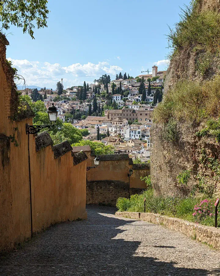 Looking down an old paved street, with a fortification on one side. In the background are the whitewashed houses of the Albaicín area of Granada.