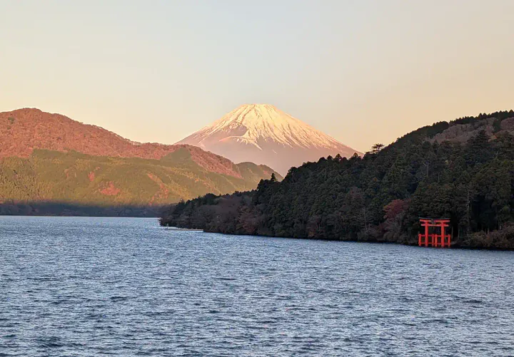 A view of Mount Fuji at sunrise - one of the benefits to staying overnight in Hakone vs taking a day trip from Tokyo. A huge, triangular, snow-capped mountain is bathed in pink light. In the foreground, there's a red shrine by the side of a lake.