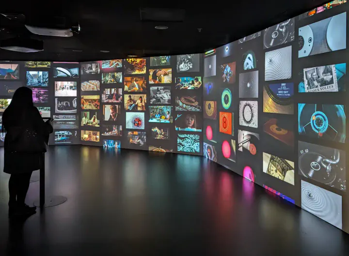 A wall in a black room, with lots of little screens all showing a different video. A woman is standing at a control screen, deciding what to watch next.