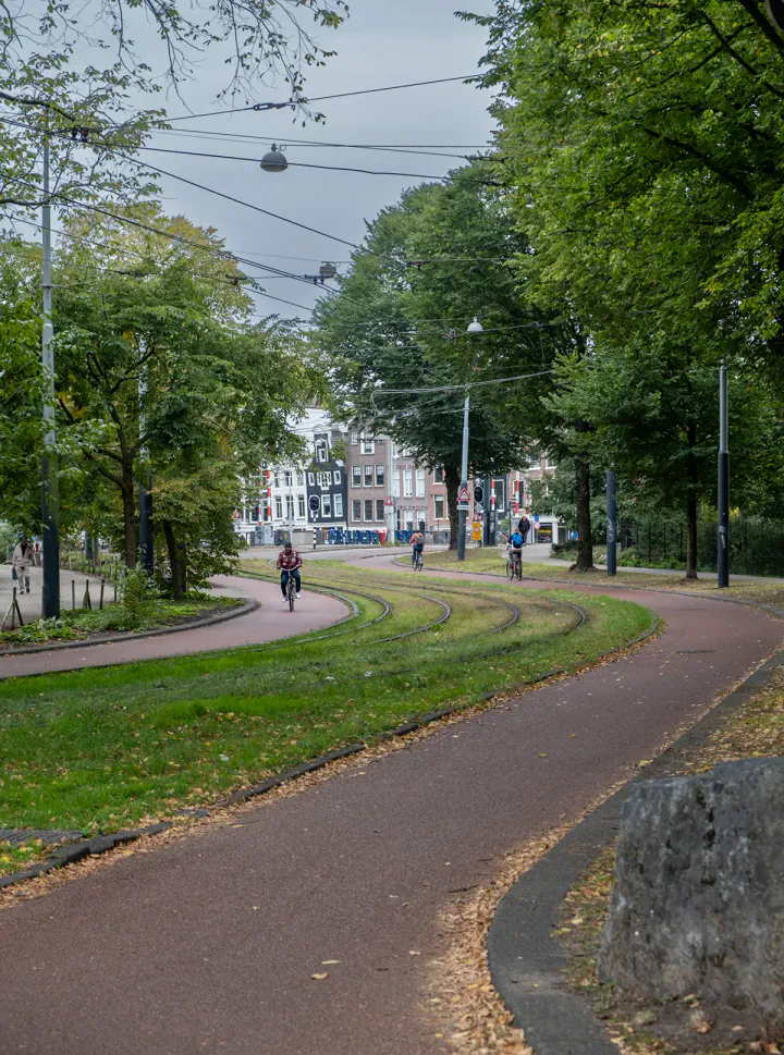A tram track curving through a park with trees. There are cyclists and typically Dutch canal houses in the background.