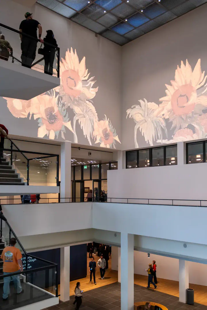 The square atrium at the Van Gogh Museum in Amsterdam. Details from Van Gogh's painting 'Sunflowers' are being projected on the white walls, while there's a staircase connecting the gallery floors. It's quite busy, with people on the stairs, on the balconies and on the ground floor.