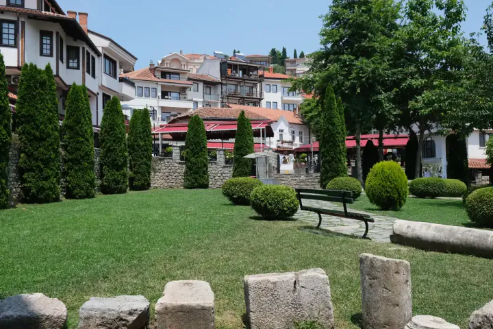 A pretty lawned garden, with benches, old stones and trees. The garden is surrounded by white houses.