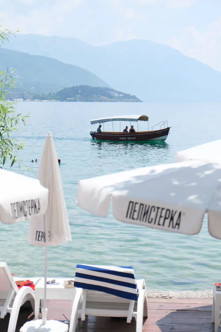 A boat sailing on a lake on a sunny day, with sun loungers and umbrellas in the foreground