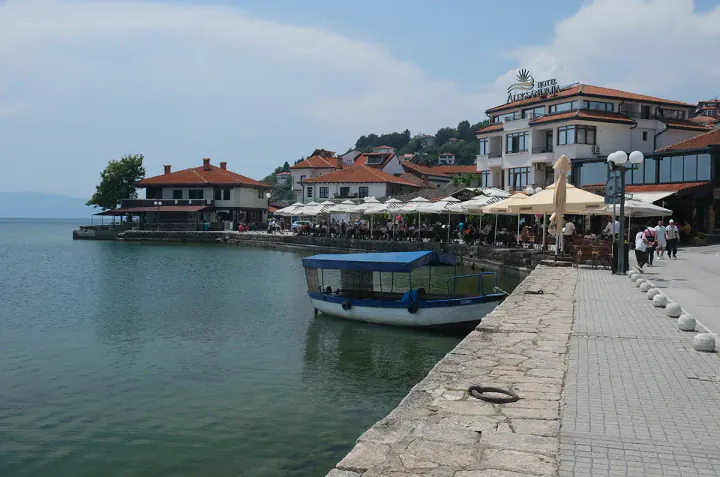 Lakeside restaurants with tables and chairs on a promenade. A little boat is moored on the quay.