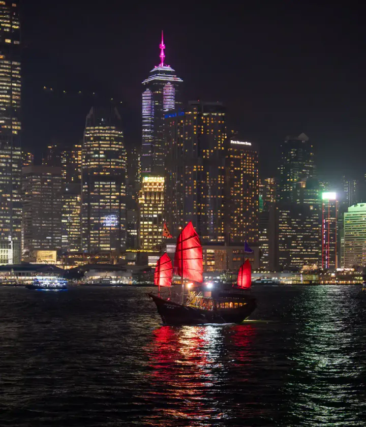 A night scene of a traditional sailing ship with red sails crossing Hong Kong's Victoria Harbour in front of skyscrapers
