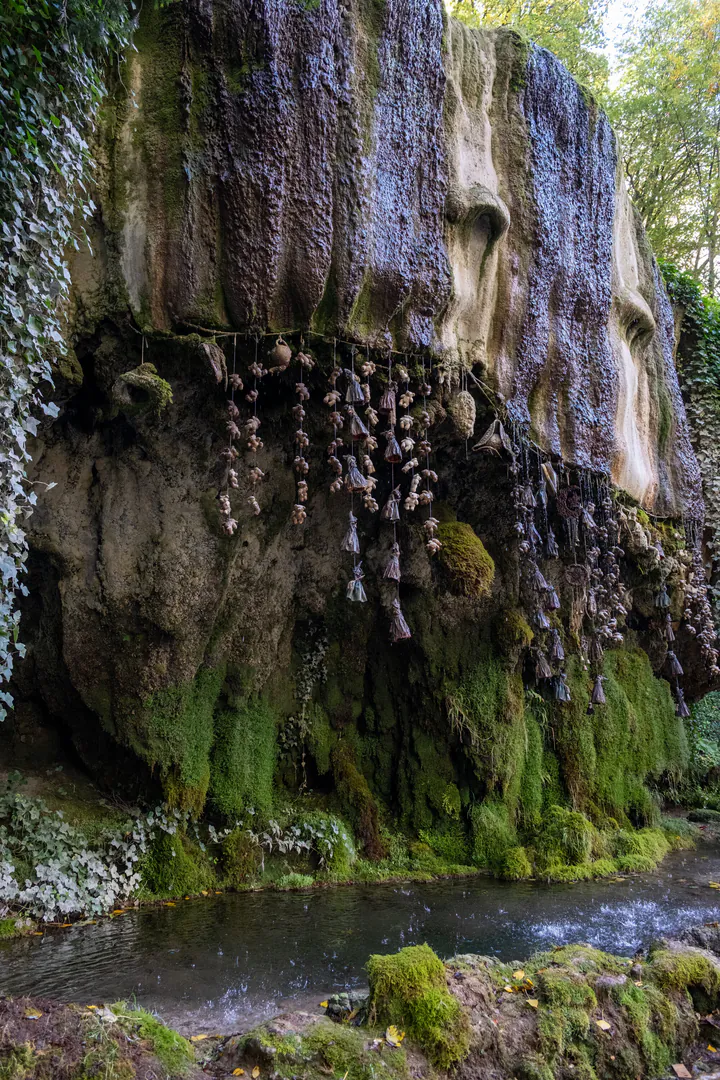A stone outcrop with water flowing over it into a well pool below. There are items including a hat, teddies and witch figurines hanging from the rocky outcrop.