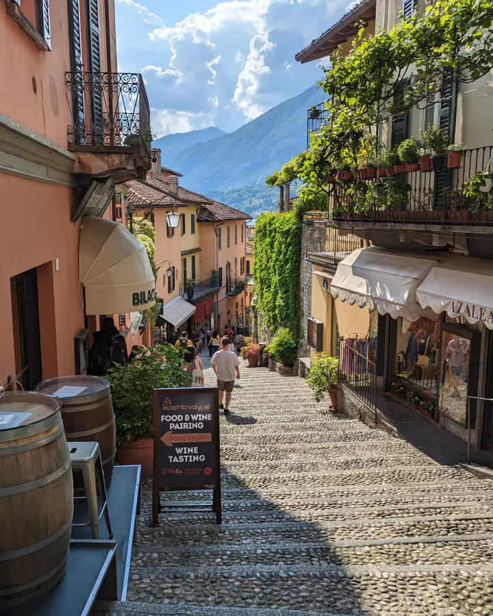 A picturesque village street in Bellagio. A cobbled street leads downhill, lined with pink and yellow buildings.