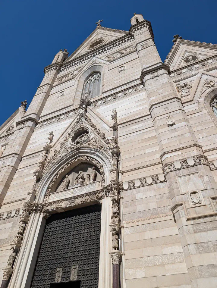 The exterior of the Naples Duomo. A fairly simple light stone building against a blue sky.