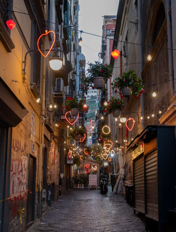 A narrow street in Naples' Spanish Quarter in the evening. There are neon hearts hanging above.