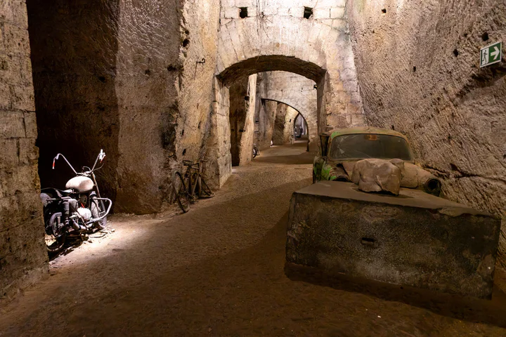 An underground tunnel with high ceilings and archways. There's an abandoned motorbike and old car.