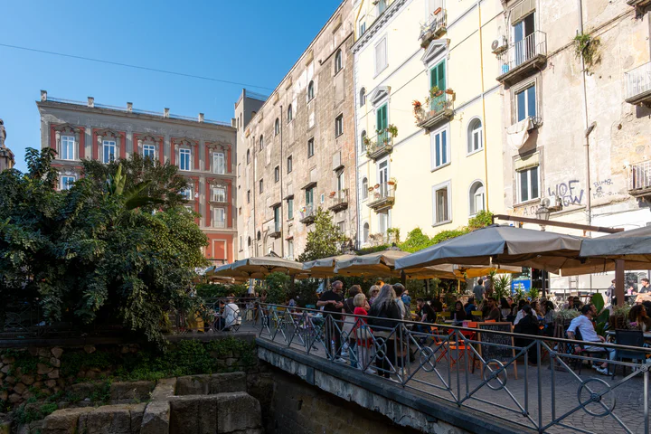 A busy city square lined with bars. There are ancient ruins in a hole in the middle.