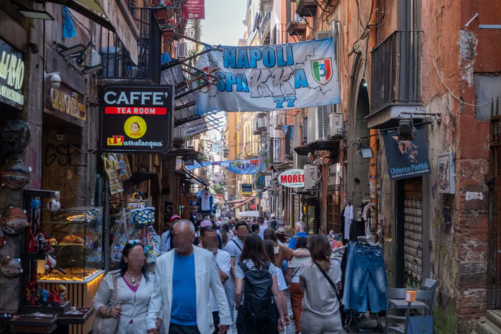 A narrow, busy street full of people. There are at least two cafes visible, plus shops, lots of signs, graffiti and football-themed banners hanging across the street.
