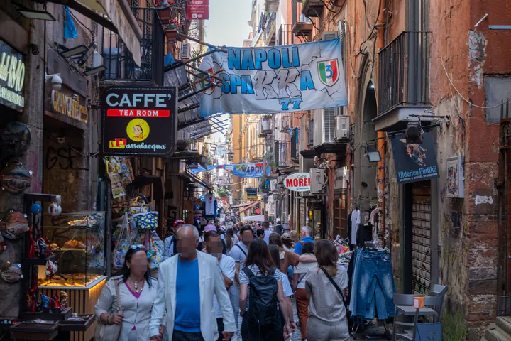 A narrow, busy street full of people. There are at least two cafes visible, plus shops, lots of signs, graffiti and football-themed banners hanging across the street.