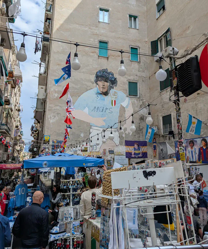 A giant mural of footballer Maradona on an apartment block's wall, with stalls selling Maradona merch on the street below.