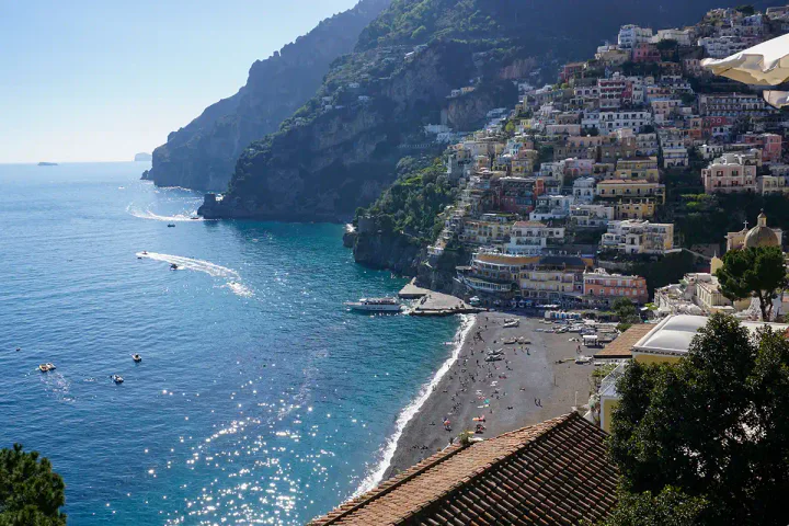 A view over Positano on the Amalfi coast. There's a pebble beach at the bottom of the cliff, with boats in the water.