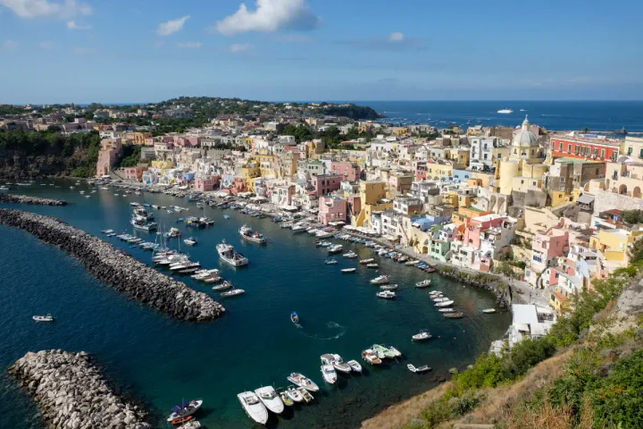A beautiful view over a curving harbour with pastel-coloured houses and a lemon-coloured church. There are boats bobbing in the harbour.