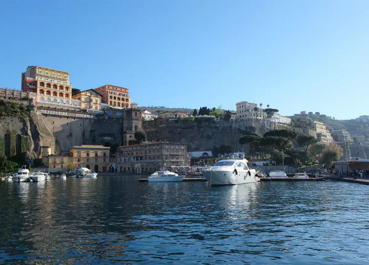 A harbour at the bottom of a cliff with a private yacht moored up. At the top of the cliff there are grand hotels.
