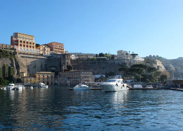 A harbour at the bottom of a cliff with a private yacht moored up. At the top of the cliff there are grand hotels.