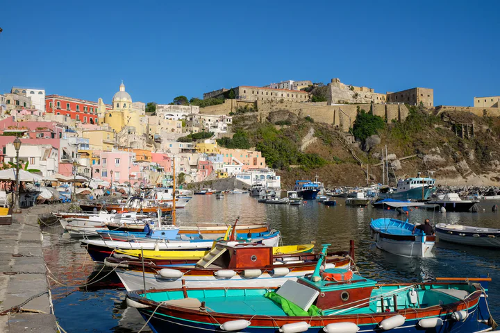 Colourful boats in the harbour of a fishing village with pastel-painted houses. There's a large stone castle on the cliff behind.