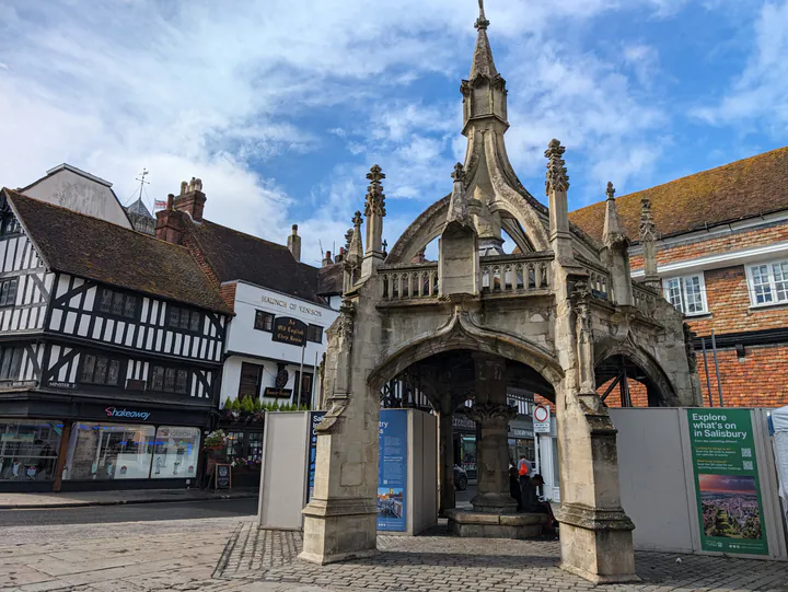 An ornate, hexagonal building with a spire on top. There are timber-framed buildings in the background.