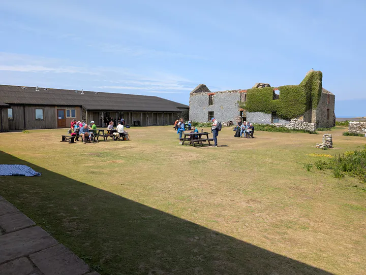 A large grassy courtyard with an abandoned stone house on one side, a low building on another side and picnic tables in the middle