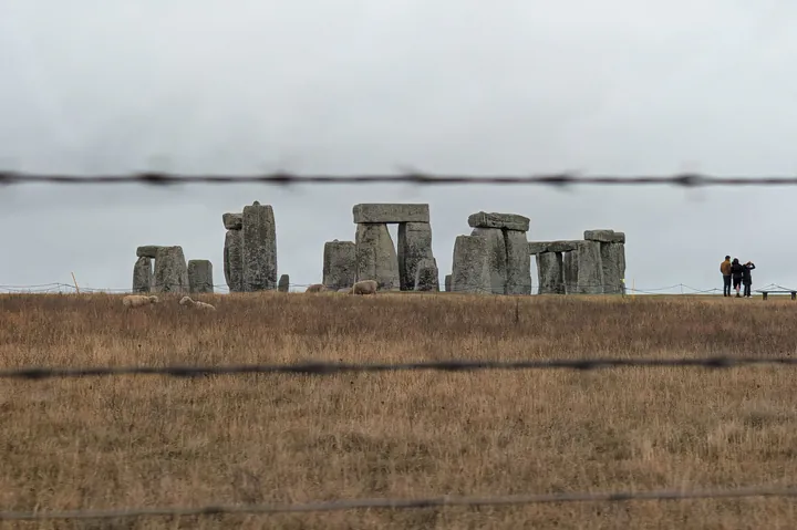 A view of Stonehenge from the A303 road. There's a field in front, and some barbed wire, but Stonehenge is clearly visible.