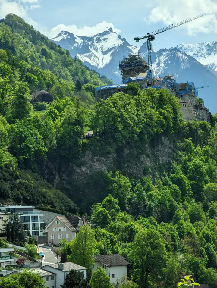 A steep cliff, covered in trees, with Vaduz Castle (Schloss Vaduz) at the top. There are snow-covered mountains in the background.