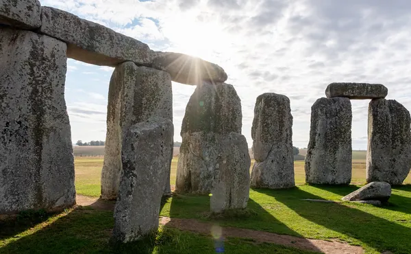 A view looking out from one of the stone circles at Stonehenge. It's the morning and there's a sunburst and long shadows.