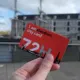 A hand holding a red I amsterdam city card 72 hours in front of an old building and sailing ship at the Maritime Museum, Amsterdam