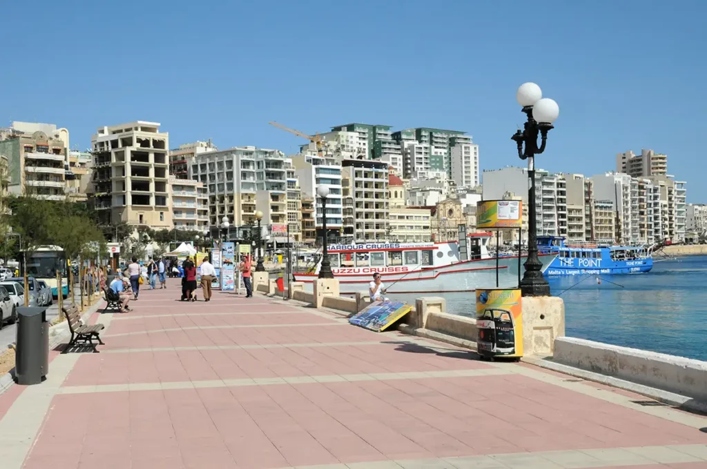 A wide seafront promenade in Sliema, Malta, with high rise buildings behind and tour boats waiting at the quay.