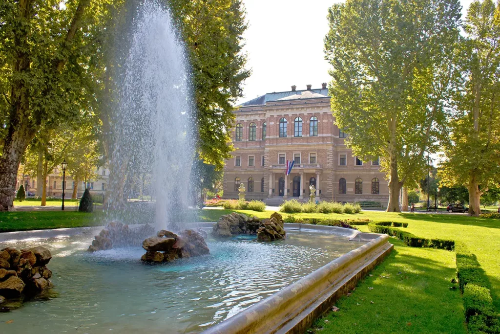 Zrinjevac Park in Zagreb, Croatia. A pool with fountains in a green city park, with a large, grand building in the background.