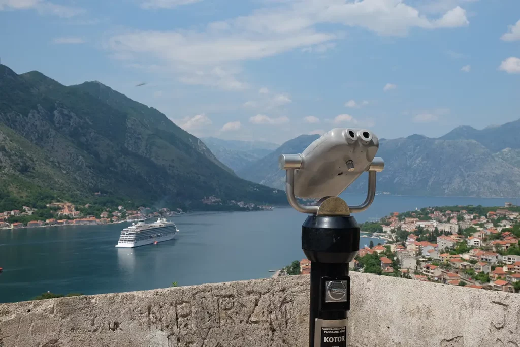 BInoculars at a viewpoint above the Bay of Kotor. There are mountains surrounding a narrow fjord and a cruise ship making its way out to sea.