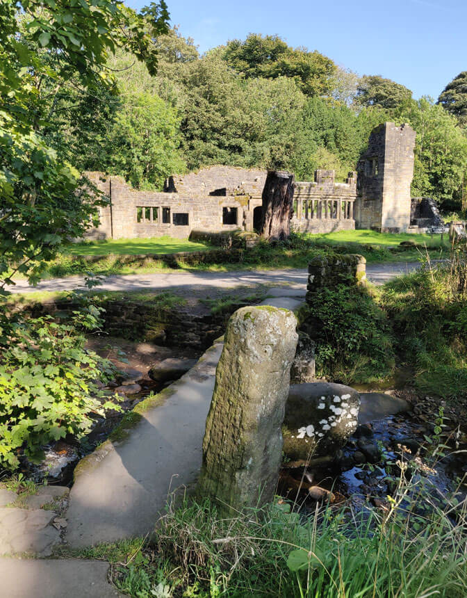 Crossing the Clapper Bridge towards the ruins of Wycoller Hall. A simple stone slab bridge crosses a narrow river. On the other side of the river are the ruins of Wycoller Hall, a large old house.