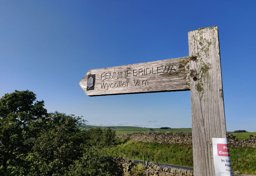 The Pennine Bridleway runs through Wycoller. A wooden sign reading Pennine Bridleway and pointing to Wycoller half a mile away.