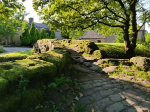 The old Pack Horse Bridge at Wycoller. A narrow, uneven, moss-covered bridge crosses a small river.