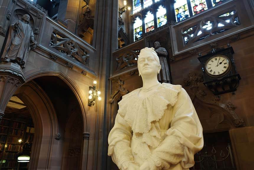 A white marble statue of Enriqueta Rylands in the reading room of the library she built. There are stone arches, statues, books and a stained glass window behind her.