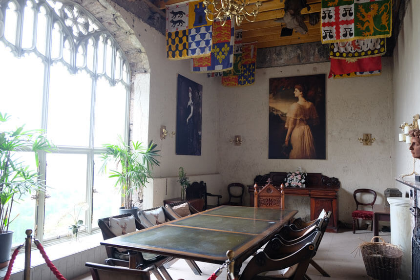 The old chapel is one of the few indoor parts of Gwrych Castle that you can visit. A high-ceilinged room inside the castle, with a large window. In the middle of the room there's a large wooden dining table surrounded by chairs. At the end of the room there's a picture of a woman wearing a white dress. There are brightly-coloured heraldic banners flying from the beams on the ceiling. 