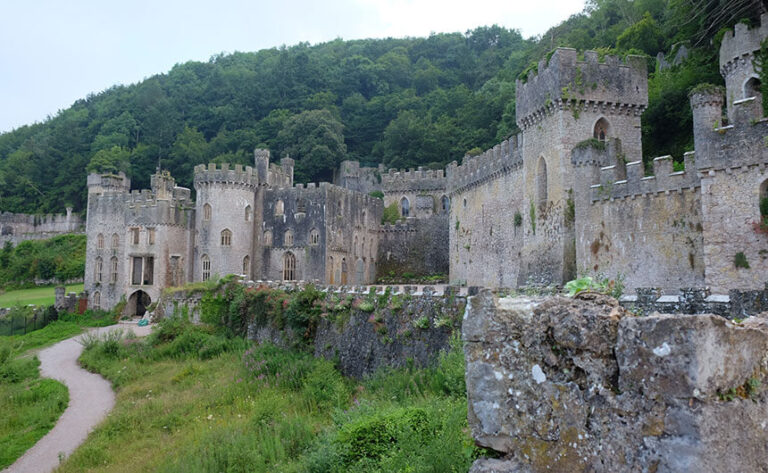 The outside of Gwrych Castle in North Wales. A ruined, roofless but very grand castle in grey stone with gothic towers spreads across a forested Welsh hillside.