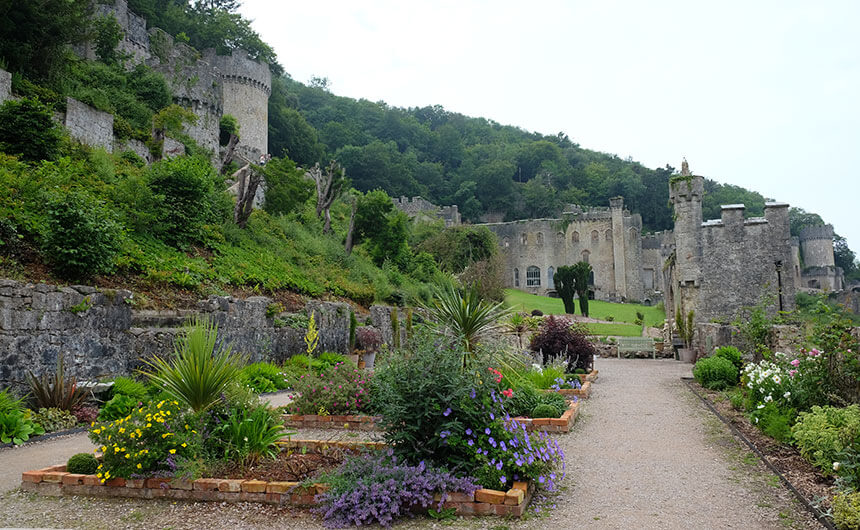 The gardens at Gwrych Castle in North Wales. Borders full of plants with yellow, purple, white and pink flowers and a green grass lawn are laid out in front of a gothic stone castle.